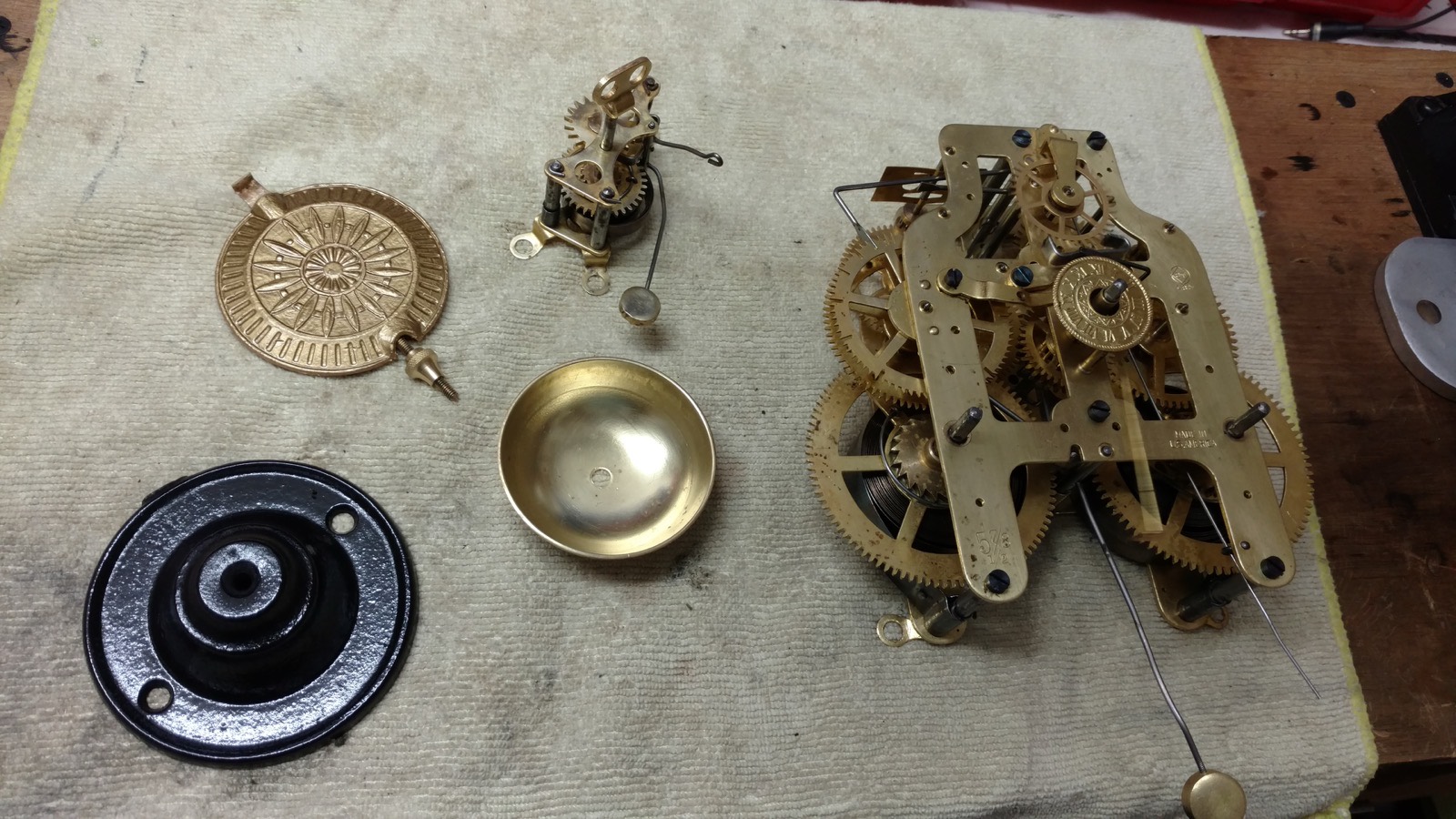 A clock movement completely disassembled on the bench — plates, wheels, mainspring, bell and pendulum bob laid out on a towel