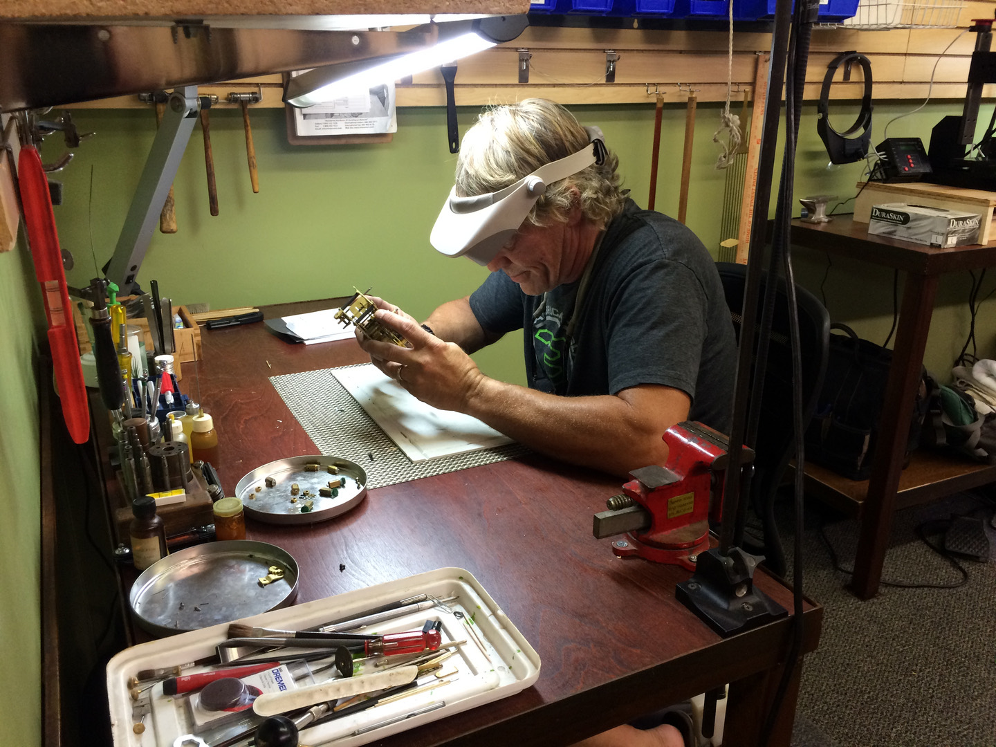 A clocksmith at the Gesswein's bench wearing a magnifier visor, inspecting a brass clock movement under the bench lamp