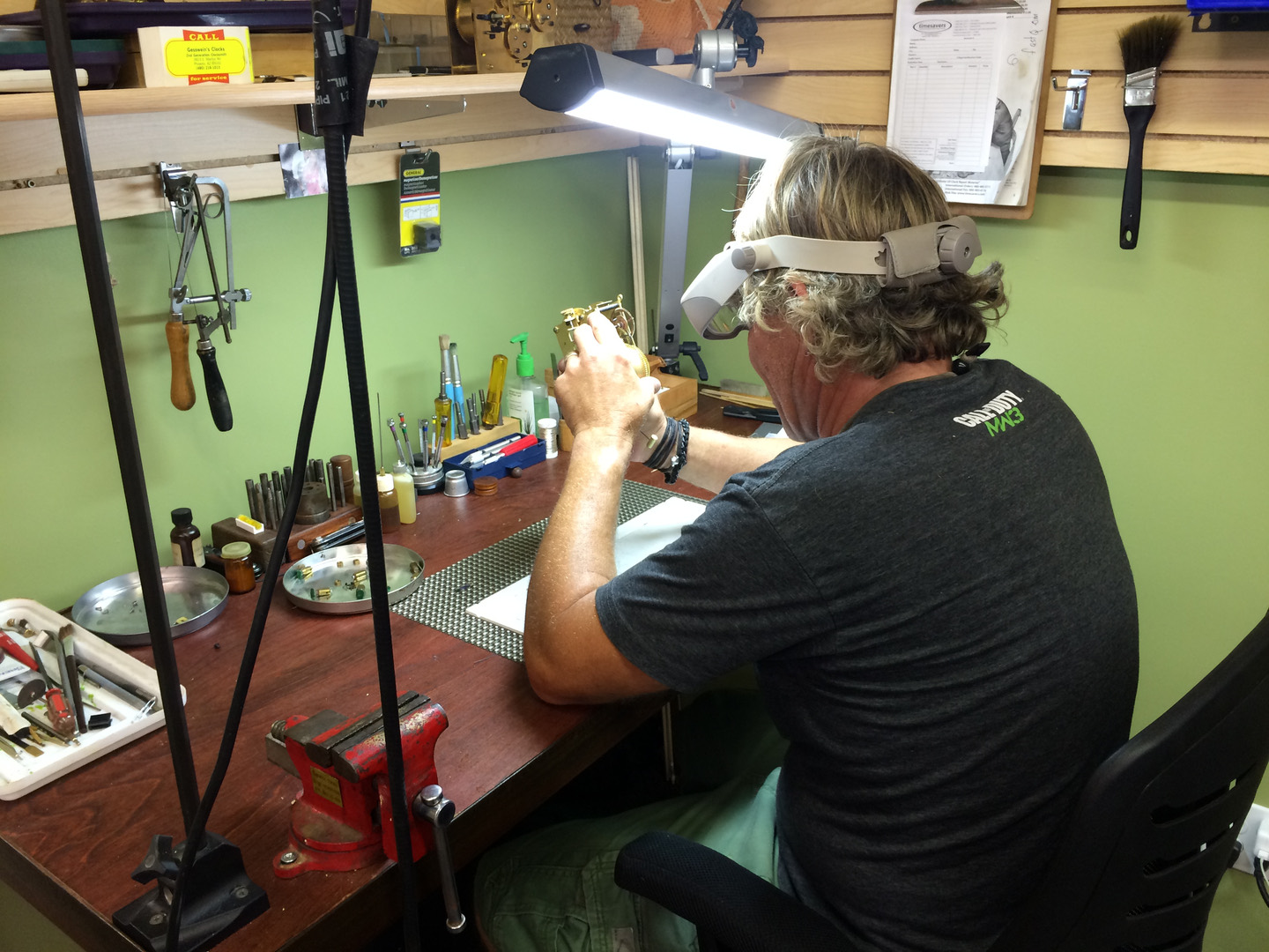 At the Gesswein's bench — a clocksmith in a visor leaning over a movement, a &lsquo;Gesswein&rsquo;s Clocks — for service&rsquo; card on the bench