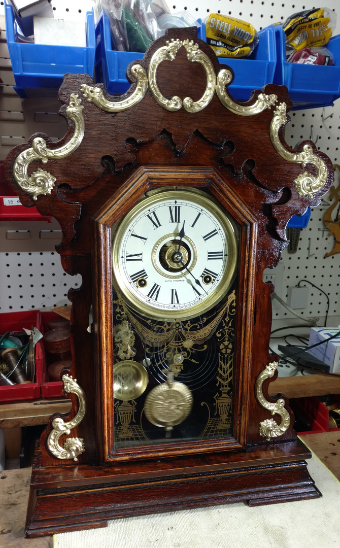 A fully restored Seth Thomas gingerbread parlor clock — carved oak case, gilt ornament and original painted glass, back on the bench and running